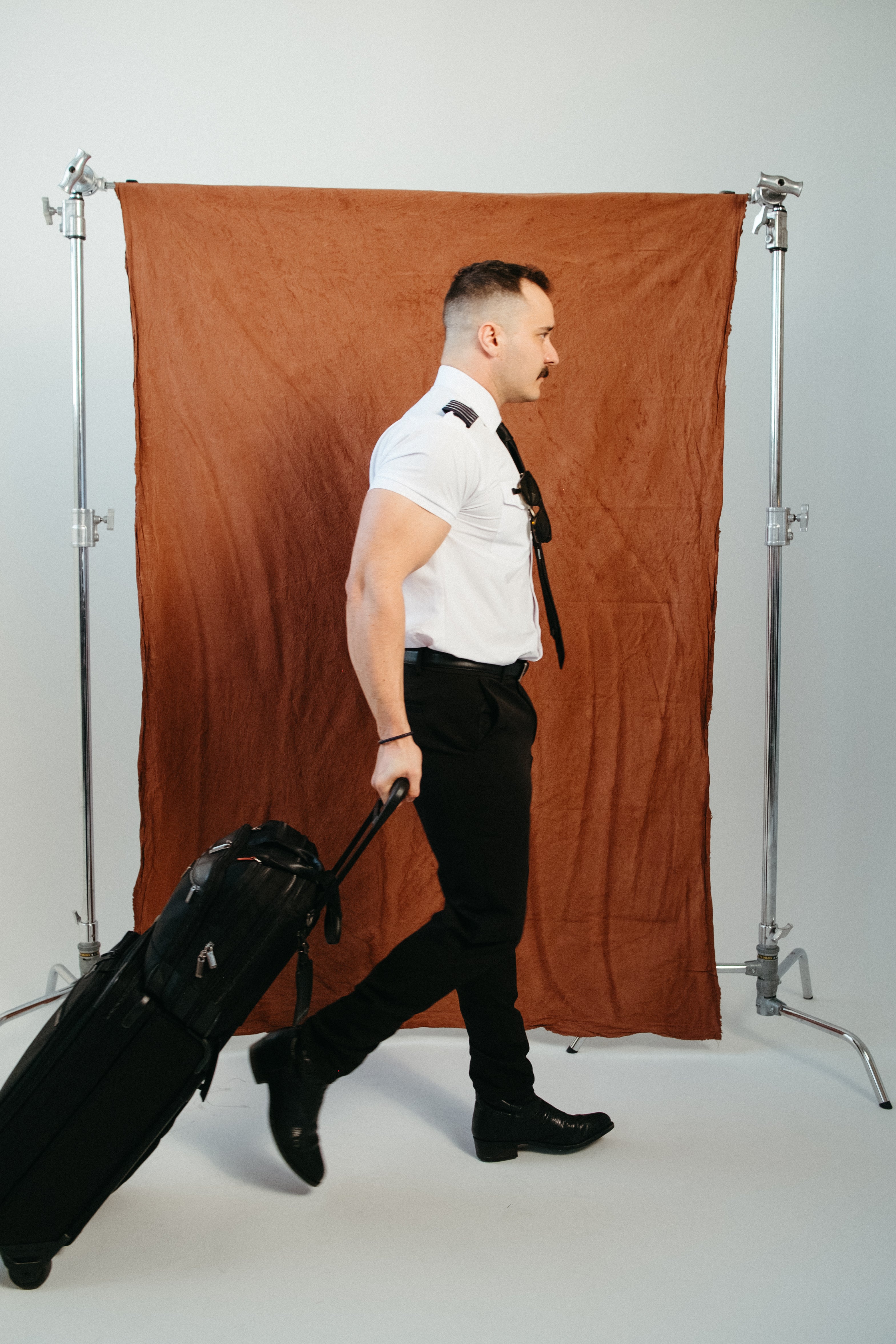 Airline pilot walking with a black rolling flight bag, wearing a white short-sleeve Flight pilot shirt with epaulettes and black tie against a rust-colored backdrop